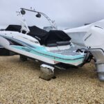 White Monterey speedboat with turquoise stripe on a trailer, Mercury outboard motor, covered seating, parked on gravel near other boats.