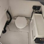 Small boat head with a white toilet, hoses, and an electrical panel in a compact cabin.