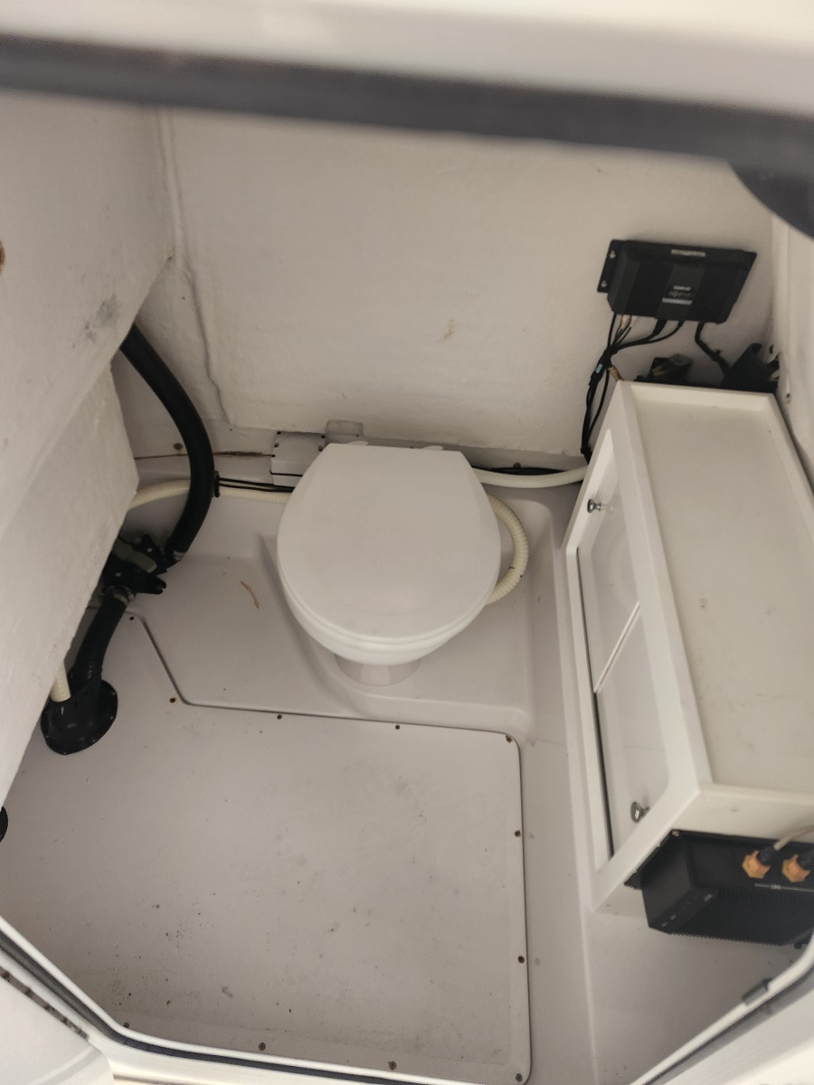 Small boat head with a white toilet, hoses, and an electrical panel in a compact cabin.