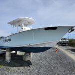 White and blue sport boat named 'CINSAN' propped on concrete blocks in a boatyard with gravel, other boats, and an orange traffic cone nearby under a clear blue sky