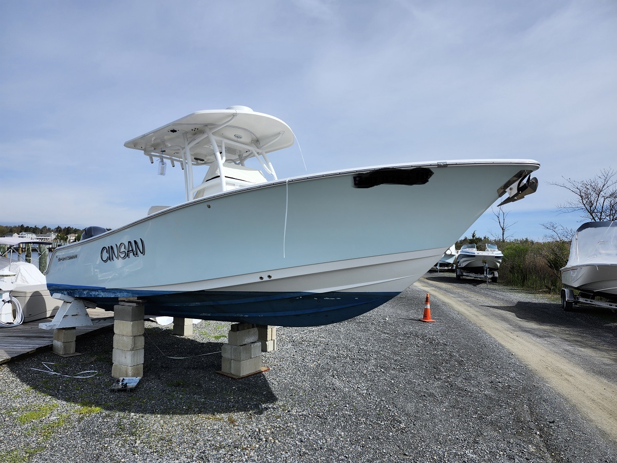 White and blue sport boat named 'CINSAN' propped on concrete blocks in a boatyard with gravel, other boats, and an orange traffic cone nearby under a clear blue sky