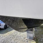 Underside of a white boat hull supported by concrete blocks on a gravel surface, with a silver car in the background.