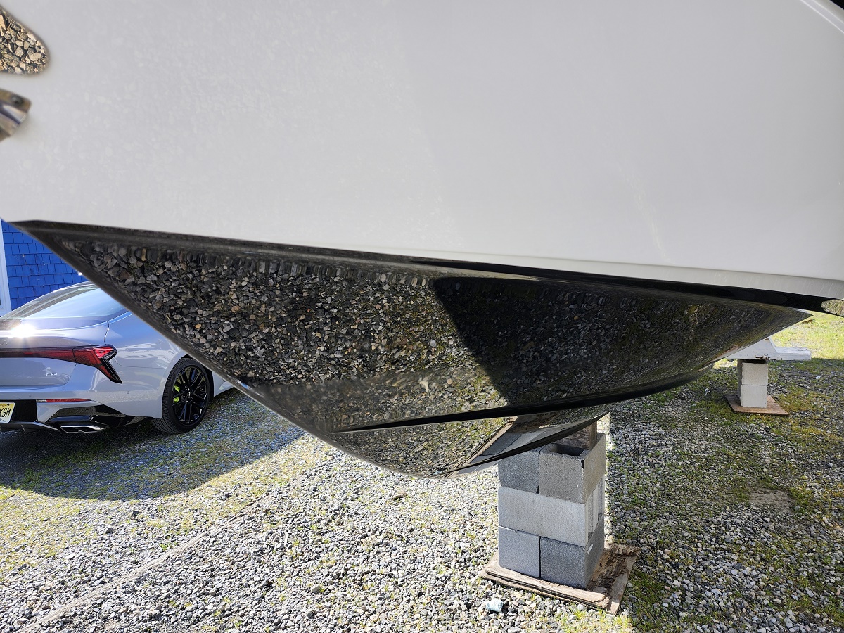 Underside of a white boat hull supported by concrete blocks on a gravel surface, with a silver car in the background.
