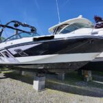 White and black Monterey speedboat on blocks in a boatyard under a bright blue sky, with a wakeboard tower visible on the left.