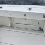 White boat cockpit scene showing a closed storage hatch, stainless support rod, and a chrome handrail; water is visible in the background.