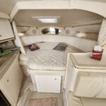 Cream-colored yacht cabin with a curved bed, quilted bedding, and built-in storage; a small galley to the left and tissue box on the counter.