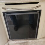 Compact under-counter RV refrigerator with a dark glass door and chrome handle marked Norcold in a beige cabinet.