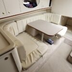 Cream-colored U-shaped seating around a rectangular table in a boat cabin, with overhead cabinets and a round porthole above.