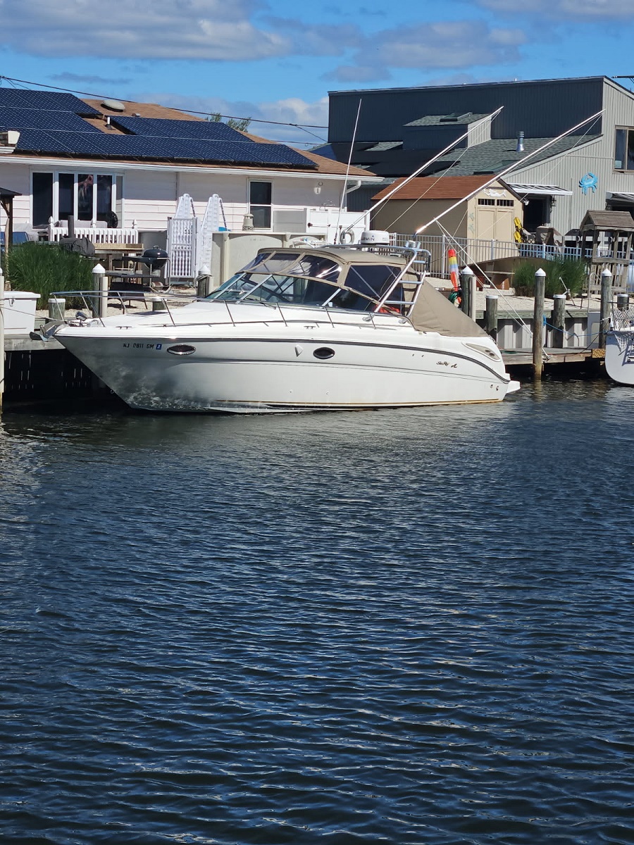 White motorboat docked at a marina beside a wooden dock, with houses and solar panels in the background under a blue sky.
