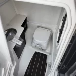 Compact marine toilet (Dometic) in a small white head, with a sink and countertop to the left and a dark floor mat nearby.