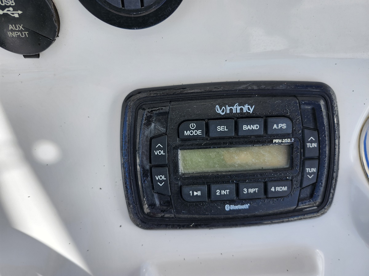 Marine stereo head unit on a white boat console, Infinity brand with Bluetooth label and numbered control buttons (volume, tune) and an LCD display.