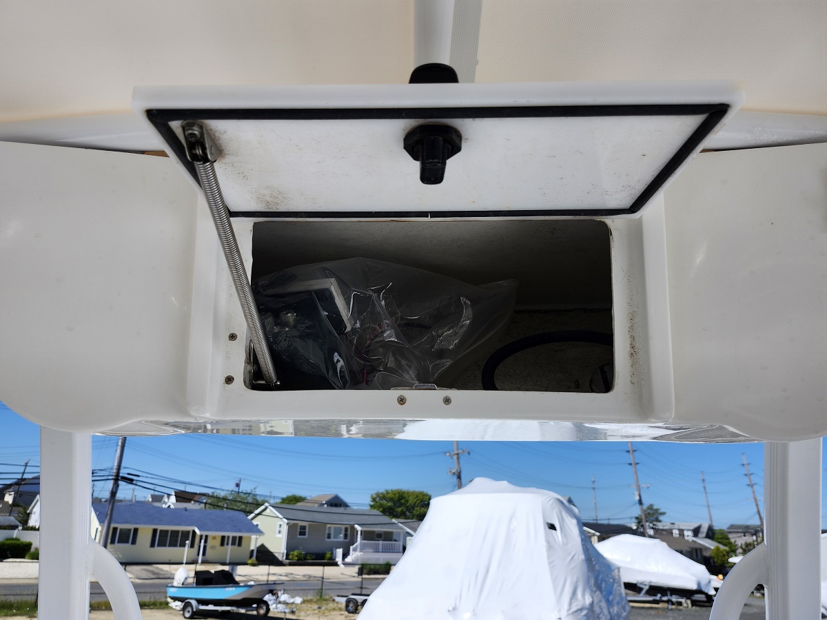 Open hatch on a white boat showing a cluttered storage compartment with a plastic bag inside, outdoors at a marina.