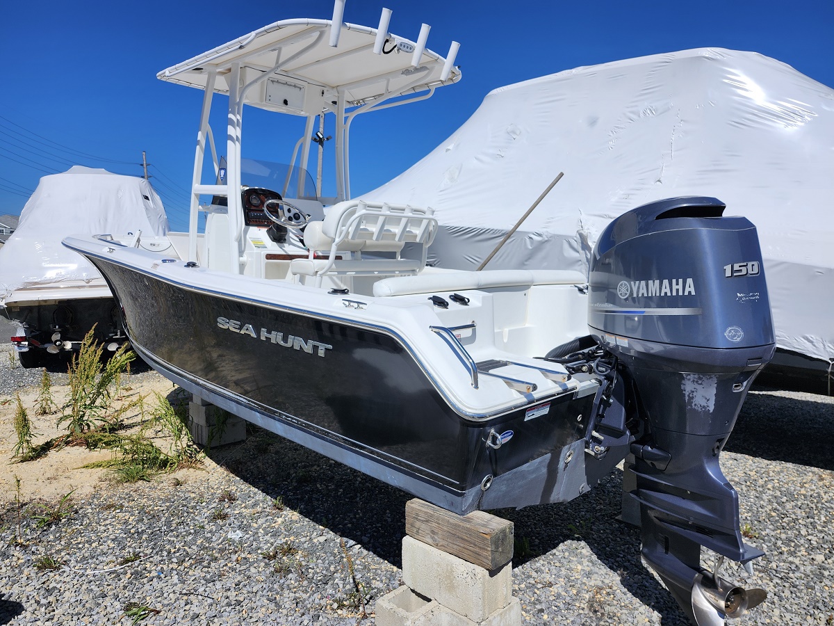 Black Sea Hunt boat on concrete blocks in a gravel lot, with a white-console cockpit and a Yamaha 150 outboard motor attached to the stern under a clear blue sky.