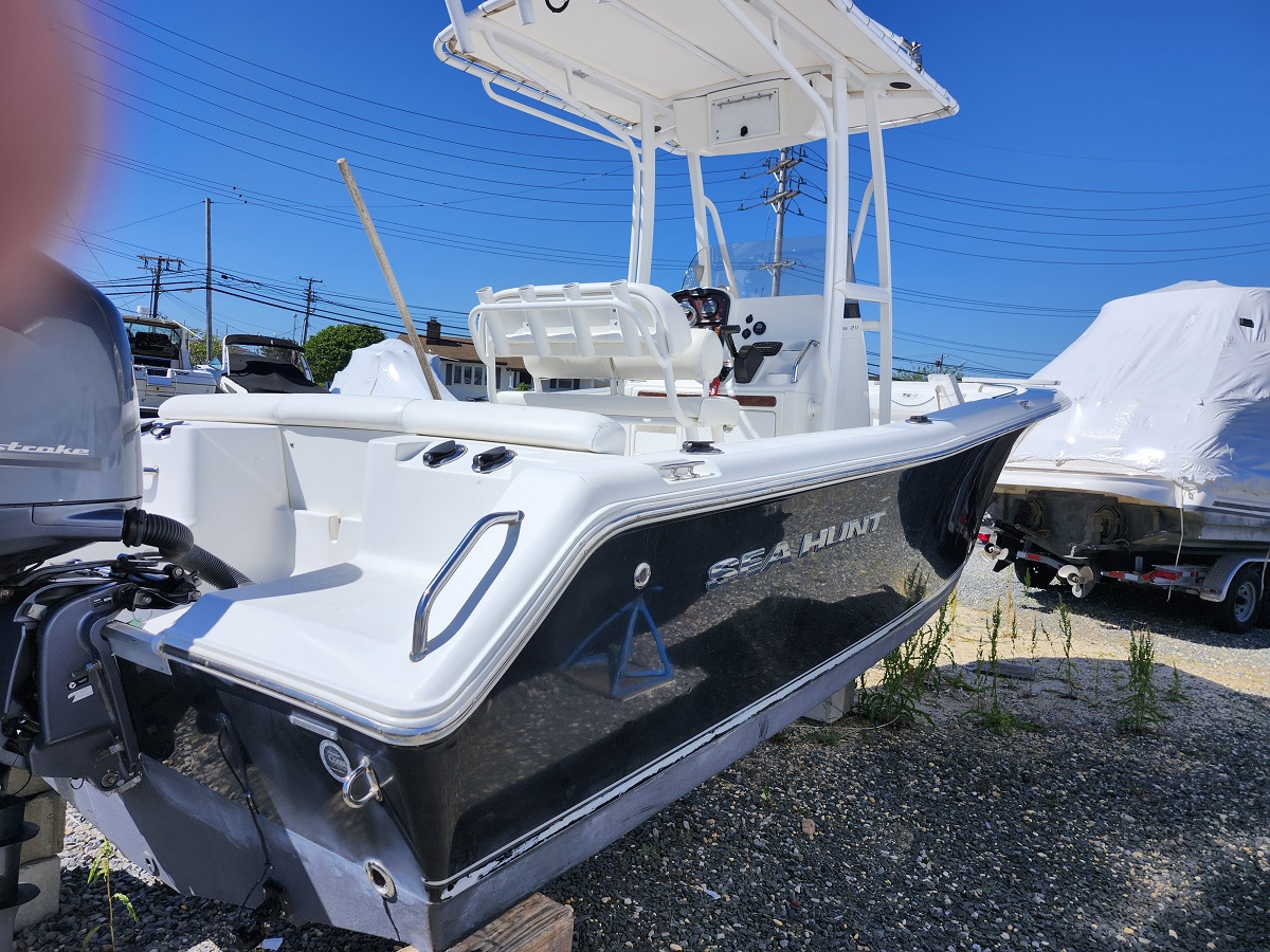 Three-quarter front view of a white-and-black Sea Hunt boat on a trailer in a boatyard under a clear blue sky, with an outboard motor visible on the left.