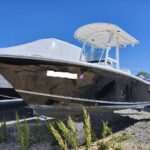 Black motorboat on concrete blocks in a gravel lot, with a white T-top and bow cover, under a clear blue sky.