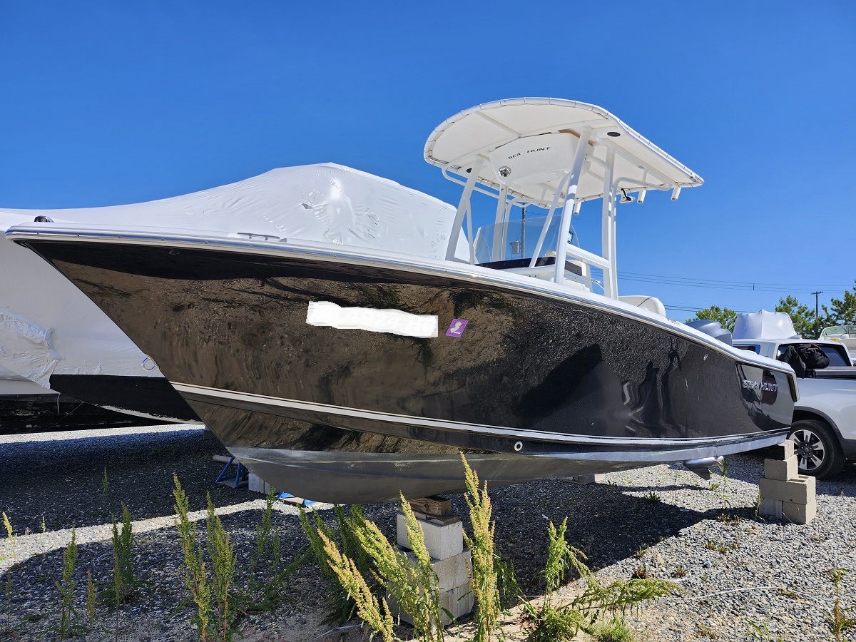 Black powerboat on concrete blocks in a gravel lot, white T-top and center console visible under a blue sky