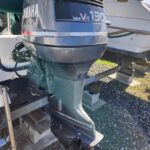 Outboard motor attached to the stern of a boat, Yamaha V8 130 engine, mounted on a stand with another boat in the background on a gravel lot.