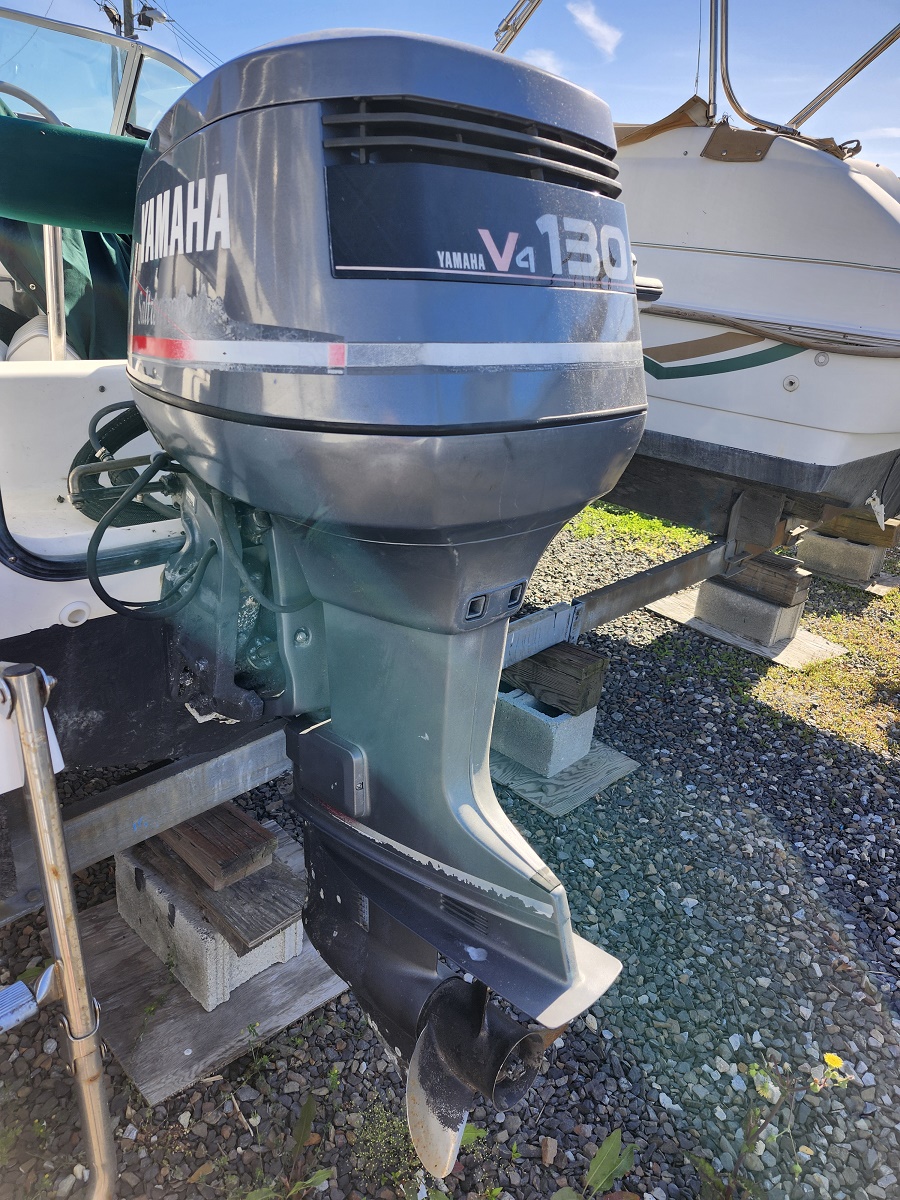 Outboard motor attached to the stern of a boat, Yamaha V8 130 engine, mounted on a stand with another boat in the background on a gravel lot.