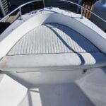 Open bow of a white motorboat showing a curved cushioned deck pad, ribbed foredeck, and stainless railing.
