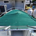 Top-down view of a boat bow with a green canvas cover and stainless railing, parked near a house and propane tanks in an outdoor yard.
