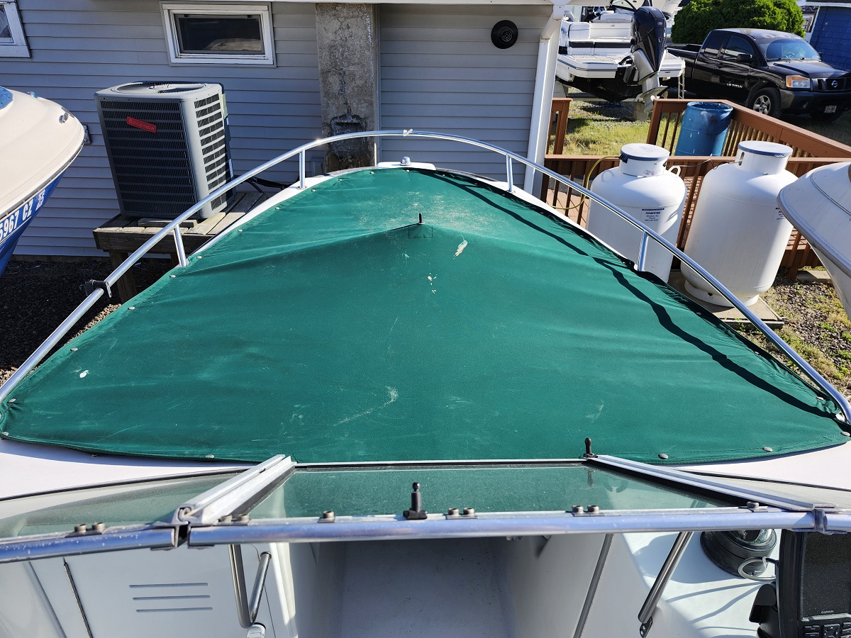 Top-down view of a boat bow with a green canvas cover and stainless railing, parked near a house and propane tanks in an outdoor yard.