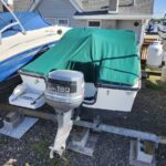Boat on a trailer with a green cover, Yamaha 130 outboard attached, parked on gravel near a house and other boats nearby in a sunny yard.