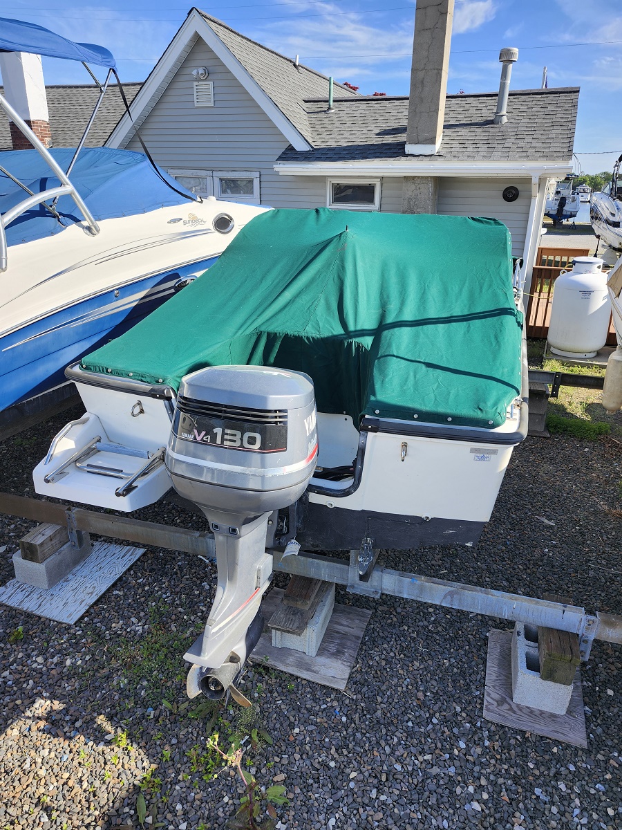 Boat on a trailer with a green cover, Yamaha 130 outboard attached, parked on gravel near a house and other boats nearby in a sunny yard.