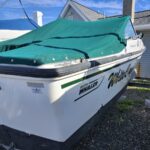 White Boston Whaler motorboat with a teal-green covered deck, parked on a trailer beside a house and gravel yard.