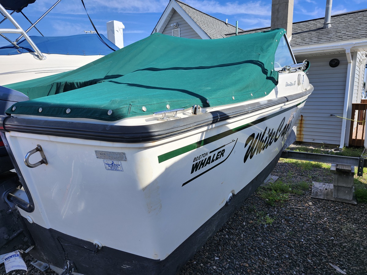 White Boston Whaler motorboat with a teal-green covered deck, parked on a trailer beside a house and gravel yard.