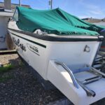 White motorboat on a trailer with a green canvas cover, parked on gravel beside a house and another boat with a Yamaha motor nearby; visible branding reads Boston Whaler on the side.