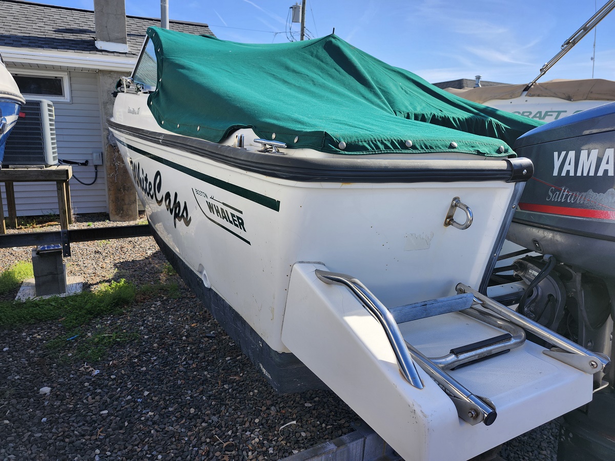 White motorboat on a trailer with a green canvas cover, parked on gravel beside a house and another boat with a Yamaha motor nearby; visible branding reads Boston Whaler on the side.