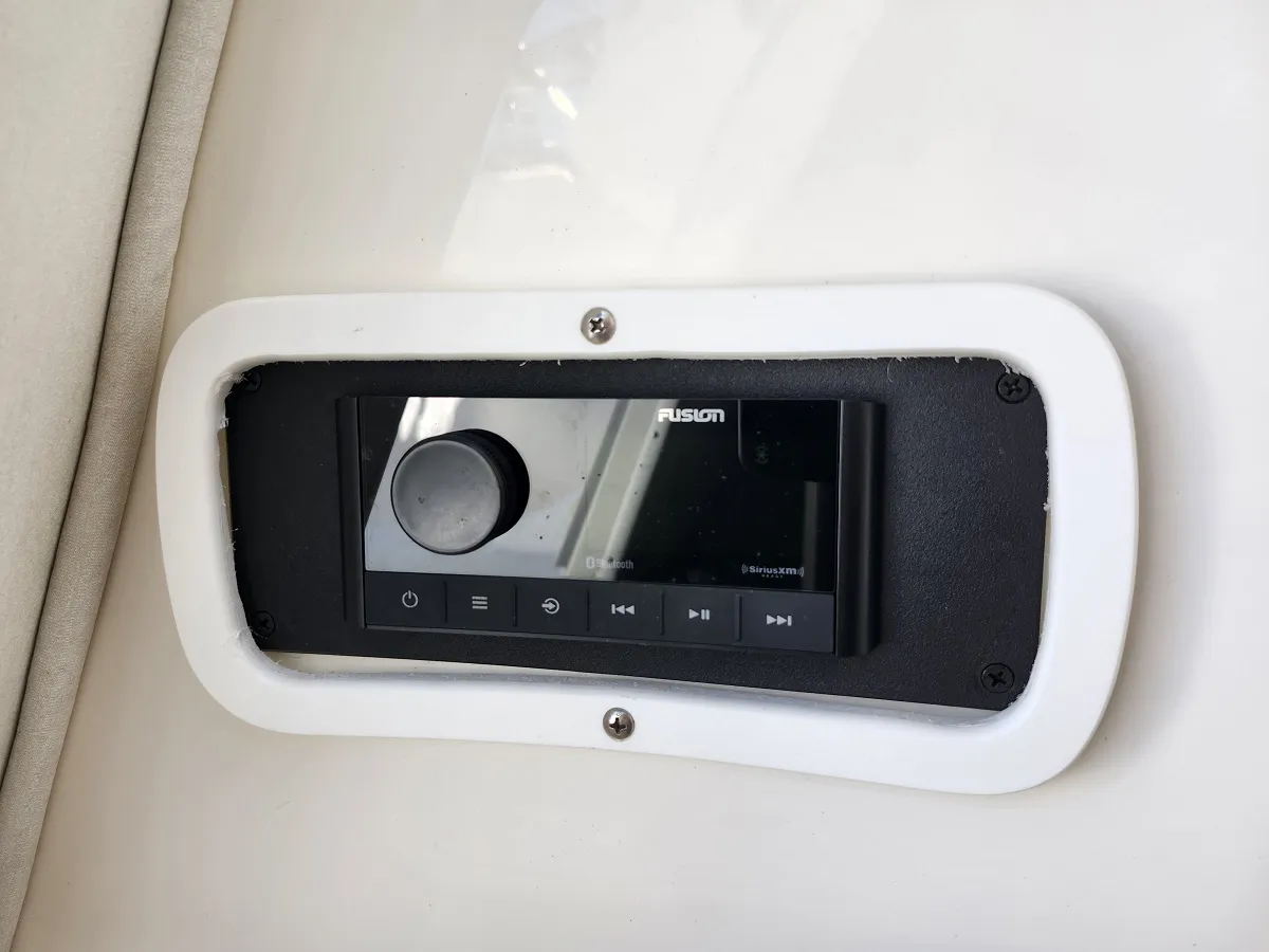 Marine stereo head unit embedded in a white panel with a black bezel; shows Fusion brand, control knob and playback buttons (power, menu, play/pause)