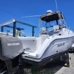 White Century motorboat elevated on concrete blocks in a boatyard, with Suzuki outboard covers and a clear blue sky above.