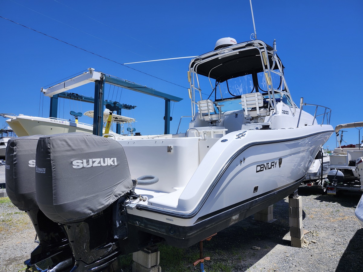 White Century motorboat elevated on concrete blocks in a boatyard, with Suzuki outboard covers and a clear blue sky above.