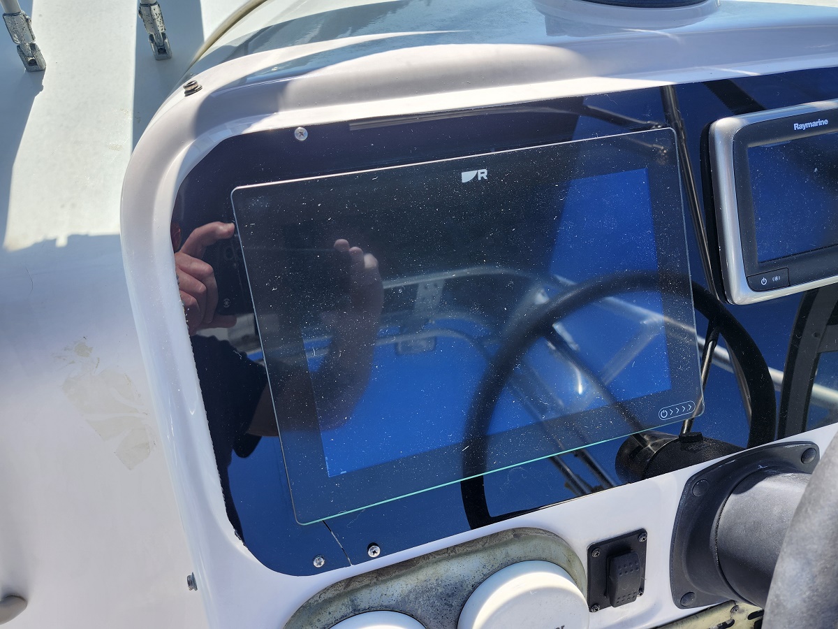 Boat helm featuring a reflective glass touchscreen display mounted at the console, with a Raymarine screen nearby in the cockpit, and a steering wheel visible in the background.