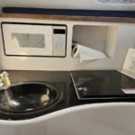 Compact galley with a built-in microwave above a black countertop, sink to the left, paper towel in an open cubby, and a smooth induction cooktop to the right.