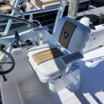 Close-up of a white boat cockpit showing steering wheel, throttle lever, and captain's chair with beige-brown upholstery in a sunny marina setting.
