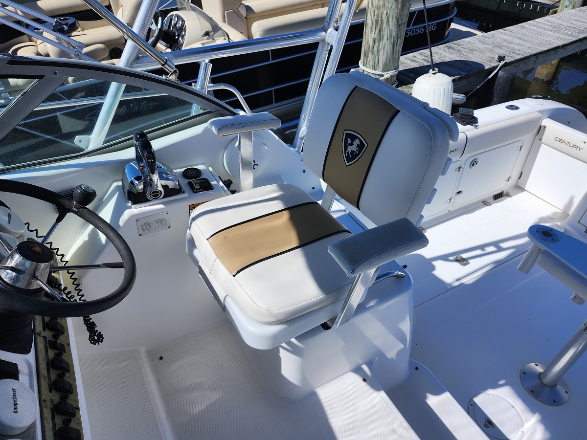 Close-up of a white boat cockpit showing steering wheel, throttle lever, and captain's chair with beige-brown upholstery in a sunny marina setting.