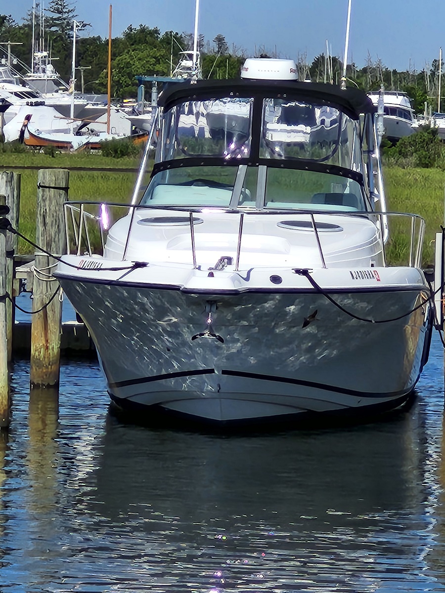 Front view of a white motorboat tied to a dock at a marina, with other boats and green shoreline in the background.