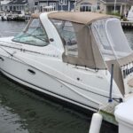 White motorboat with a tan canvas cover docked at a marina, calm water and houses in the background.