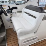 Captain's chair at the boat's helm with steering wheel, throttles, and control panel; white and gray upholstery on a teak deck