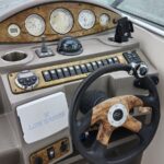 Boat helm with wood-grain dashboard, multiple gauges, and a central steering wheel with a wooden grip. Most gauges show speedometer and engine readouts.