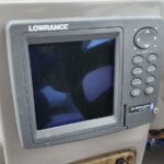 Lowrance fish finder/chartplotter mounted on boat console, screen off, with circular navigation pad and labeled buttons on the right.