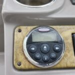 Close-up of a circular marine stereo control panel with a small display, power button labeled POWER SOURCE above, and volume/track buttons on a worn beige dashboard plate.