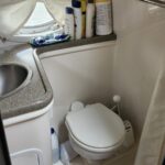 Small bathroom with a sink on a speckled countertop, row of toiletries, and a close-by toilet under a window with a curtain.