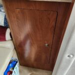 Wooden cabinet door with rounded inset panel and metal knob under a beige countertop, with a laundry basket and clutter nearby on carpeted floor.