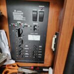 Close-up of a generator control panel mounted in a wooden cabinet, showing switches, indicator lights, and a gauge with tools (scissors, pliers) at the bottom shelf nearby.