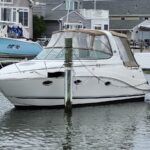 White motorboat docked at a marina with a tan canvas canopy and multiple mooring cleats, surrounded by pilings and calm water behind a row of houses.