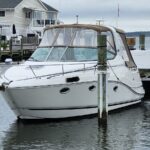 White motor yacht docked at a marina with beige canvas cover and houses along the waterfront in the background.
