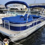 Blue pontoon boat with a blue canopy docked at a marina, white railings and houses in the background